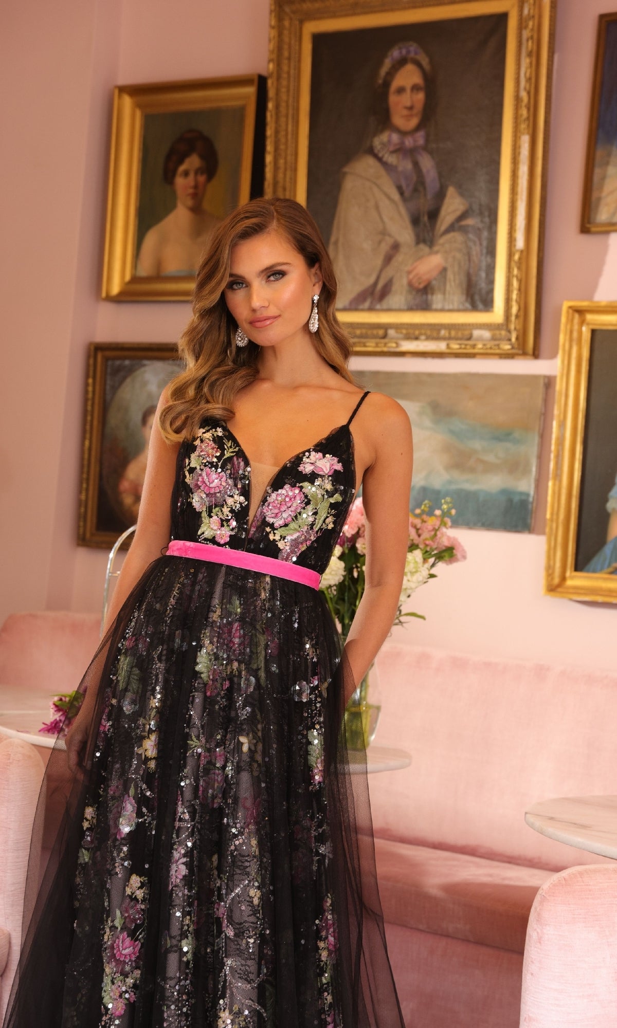 A woman in the Ava Presley 47013 long formal dress—a black V-neck ball gown with floral sequins—stands in a pink room with portraits on the wall behind her.